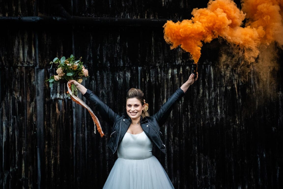 A same sex bride holding up smoke against the black of the Dutch Barn, the outside