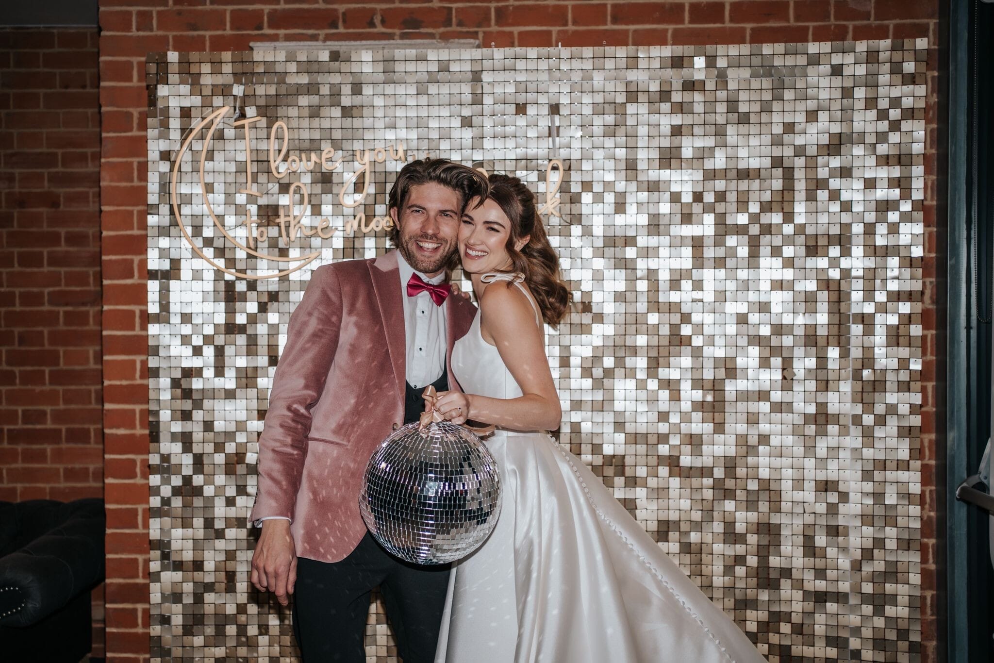 A couple stand in front of a glittering wall ready for their couple shots